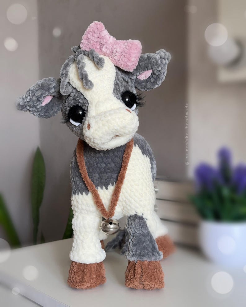 May include: A plush cow toy made of soft gray and white yarn with a pink bow on its head. The cow has large black eyes with long eyelashes, a brown collar with a silver bell, and brown hooves. The text @vareshka_handmade is visible on the side.