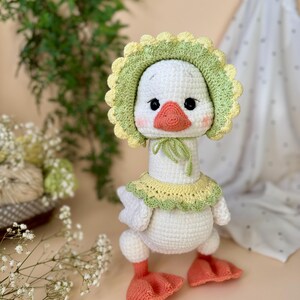May include: A handmade white crochet goose wearing a green and yellow bonnet and collar. The goose has a peach-colored beak and feet, and black eyes. The plush toy is standing upright.
