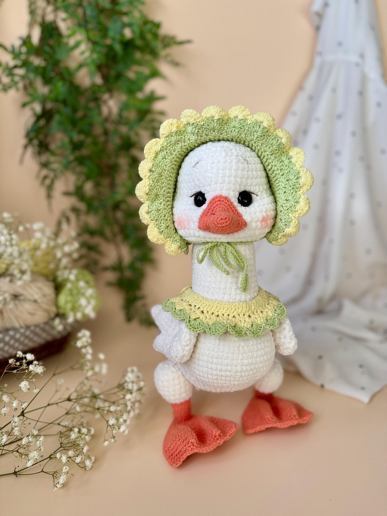 May include: A handmade white crochet goose wearing a green and yellow bonnet and collar. The goose has a peach-colored beak and feet, and black eyes. The plush toy is standing upright.