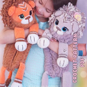 Könnte beinhalten: Zwei Plüschlöwen-Spielzeuge in Orange- und Lavendeltönen werden von einer Person gehalten. Der orangefarbene Löwe hat eine braune Mähne und der lavendelfarbene Löwe eine hellviolette Mähne. Beide haben weiße Pfoten. Der Text "CROCHET PATTERN PDF" ist sichtbar.