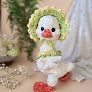 May include: A handmade white crochet goose doll with a yellow and green bonnet and collar. The goose has a peach-colored beak and feet, and black eyes. The doll is a soft, plush toy.