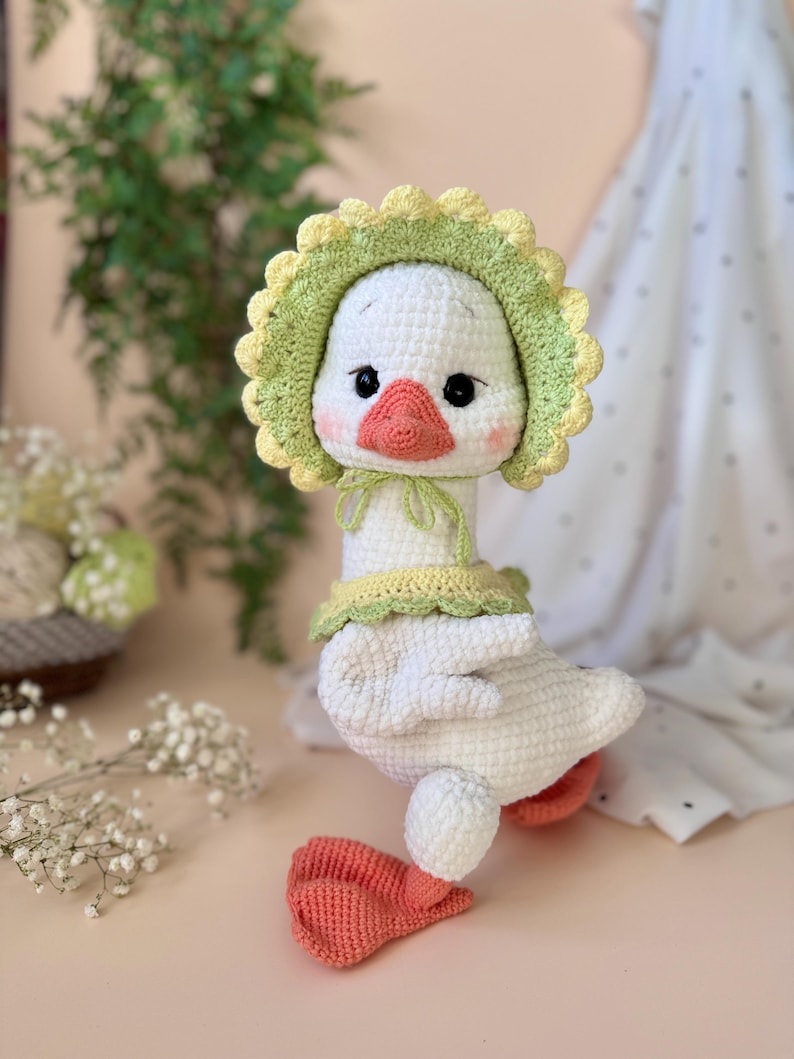 May include: A handmade white crochet goose doll with a yellow and green bonnet and collar. The goose has a peach-colored beak and feet, and black eyes. The doll is a soft, plush toy.
