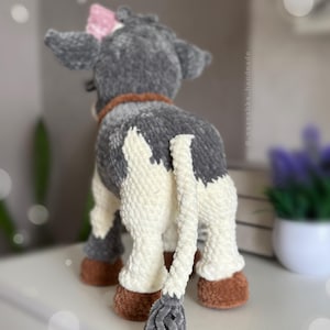 May include: A plush cow toy made of soft gray and white yarn, featuring a brown collar, brown hooves, and a small pink bow on its head. The tail is long and white with a gray tuft at the end. The text _varezhka_handmade is visible on the side.
