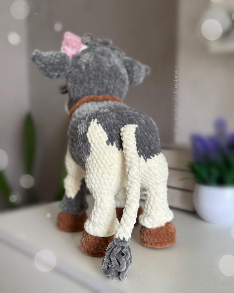 May include: A plush cow toy made of soft gray and white yarn, featuring a brown collar, brown hooves, and a small pink bow on its head. The tail is long and white with a gray tuft at the end. The text _varezhka_handmade is visible on the side.
