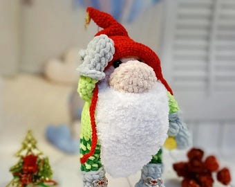 May include: A handmade crochet gnome doll with a red hat, white beard, and gray mittens and boots. The gnome has a green body with a patterned design and a red string. The doll is a festive holiday decoration.