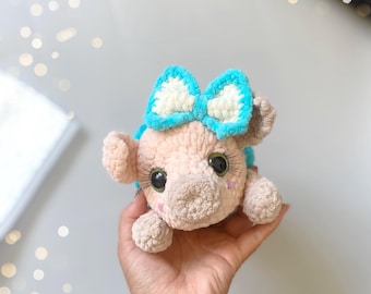 May include: A crocheted pig toy with a blue bow, brown eyes, and a pink body. The toy is being held in a hand.