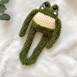 May include: A handmade green crochet frog plush toy with large black eyes and a yellow and white striped belly. The frog has long arms and legs, and is laying on a white, fluffy surface.
