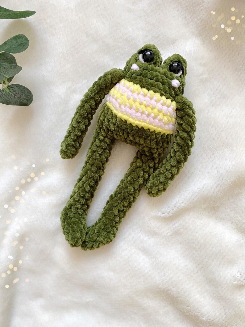 May include: A handmade green crochet frog plush toy with large black eyes and a yellow and white striped belly. The frog has long arms and legs, and is laying on a white, fluffy surface.