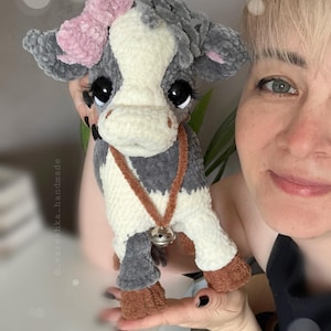 May include: A plush cow toy made of soft gray and white yarn with a pink bow on its head, large black eyes with long eyelashes, and a brown collar featuring a small silver bell. The toy has brown hooves and a textured, fuzzy appearance.