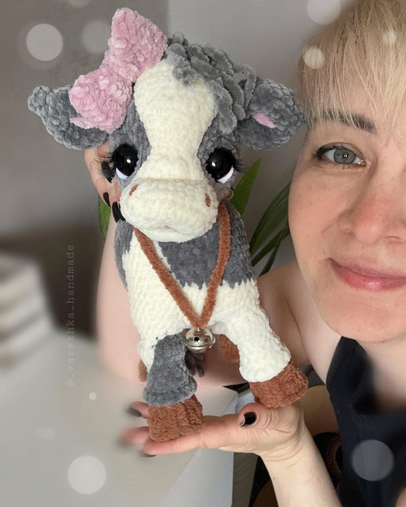 May include: A plush cow toy made of soft gray and white yarn with a pink bow on its head, large black eyes with long eyelashes, and a brown collar featuring a small silver bell. The toy has brown hooves and a textured, fuzzy appearance.