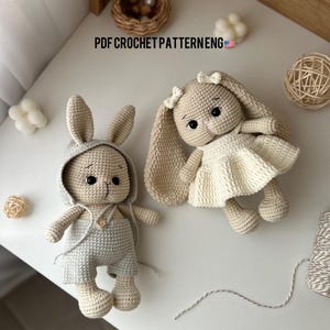 Pode incluir: Dois brinquedos de coelho de crochê, um em bege com um vestido branco e o outro em cinza com um macacão azul. Ambos os coelhos têm orelhas compridas e olhos pretos. Os brinquedos estão sentados em uma superfície branca com um fundo de madeira.