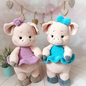 Op de afbeelding: Twee gehaakte varkens knuffels die jurken dragen. E&eacute;n varken draagt een paarse jurk met een roze strik, en de andere draagt een blauwe jurk met een blauwe strik.