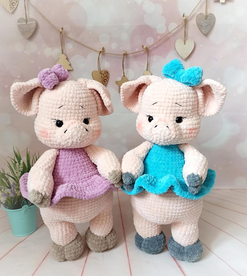 Op de afbeelding: Twee gehaakte varkens knuffels die jurken dragen. E&eacute;n varken draagt een paarse jurk met een roze strik, en de andere draagt een blauwe jurk met een blauwe strik.