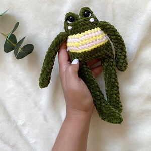 May include: A handmade green crochet frog plush toy with a yellow and pink striped belly. The frog has large black eyes and white accents. The plush toy is soft and cuddly.