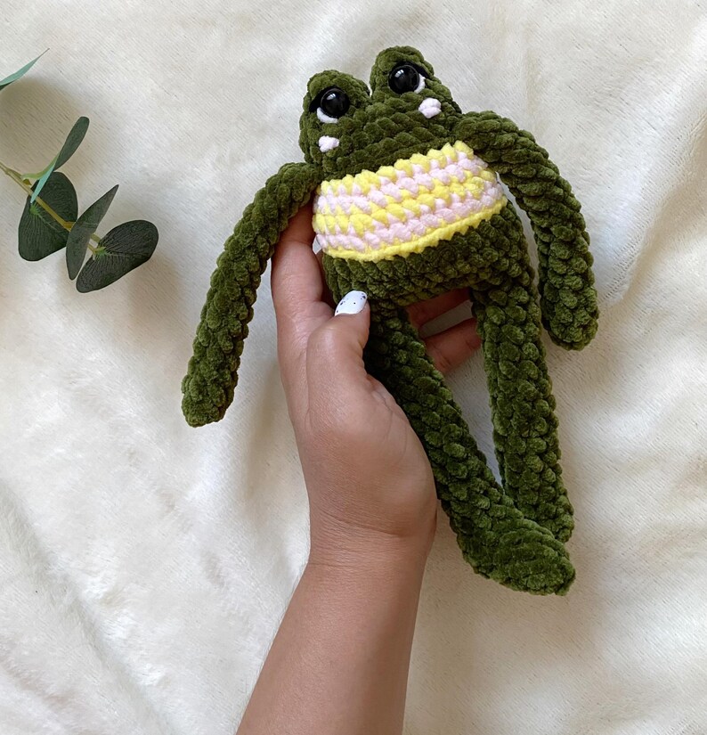 May include: A handmade green crochet frog plush toy with a yellow and pink striped belly. The frog has large black eyes and white accents. The plush toy is soft and cuddly.