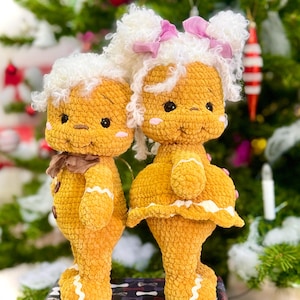 Puede incluir: Dos mu&ntilde;ecos de jengibre de crochet, uno con un lazo blanco y rosa y el otro con un lazo marr&oacute;n, de pie frente a un &aacute;rbol de Navidad.