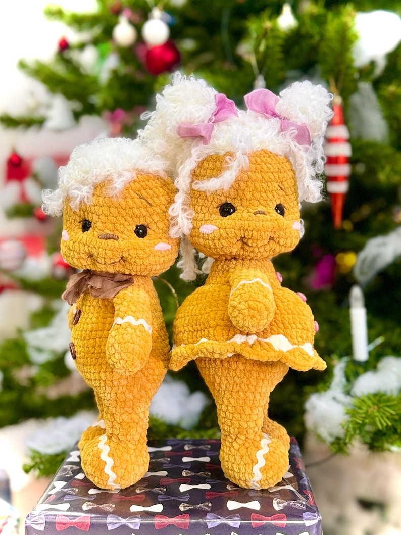Puede incluir: Dos mu&ntilde;ecos de jengibre de crochet, uno con un lazo blanco y rosa y el otro con un lazo marr&oacute;n, de pie frente a un &aacute;rbol de Navidad.
