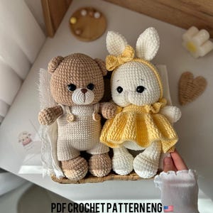 Puede incluir: Un oso de crochet marrón que lleva un atuendo de crochet blanco y un conejito de crochet amarillo que lleva un vestido de crochet blanco. Los animales de peluche están sentados en una cesta de mimbre. El texto "PDF CROCHET PATTERN ENG" es visible en la imagen.