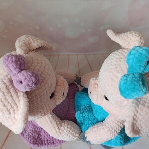Op de afbeelding: Twee gehaakte varkens knuffels, een met een paarse strik en de andere met een blauwe strik. De knuffels zijn gemaakt van zacht, pluche garen en hebben een zoete, speelse uitstraling.