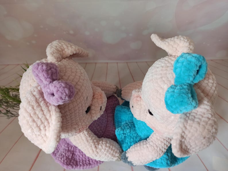 Op de afbeelding: Twee gehaakte varkens knuffels, een met een paarse strik en de andere met een blauwe strik. De knuffels zijn gemaakt van zacht, pluche garen en hebben een zoete, speelse uitstraling.