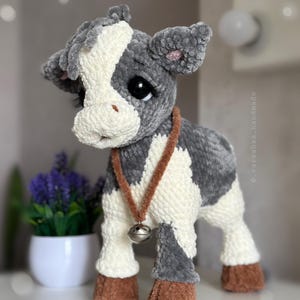 May include: A plush cow toy made of soft gray and white yarn with a textured finish. The cow features large black eyes with long lashes, brown hooves, and a brown cord necklace with a small silver-toned metal bell hanging around its neck.