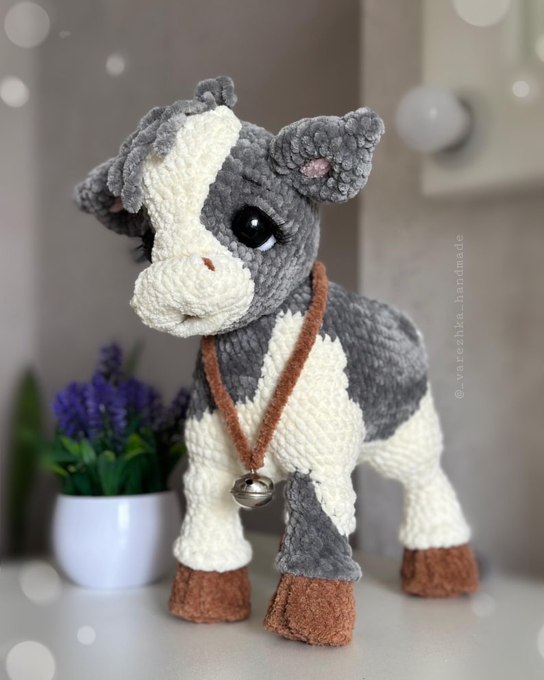 May include: A plush cow toy made of soft gray and white yarn with a textured finish. The cow features large black eyes with long lashes, brown hooves, and a brown cord necklace with a small silver-toned metal bell hanging around its neck.