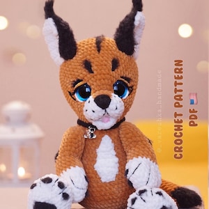 Op de afbeelding: Een gehaakt pluche speelgoed van een caracal kat. De kat heeft een bruin lichaam, witte poten met zwarte vlekken en grote blauwe ogen. Het draagt een zwarte halsband met een kleine bedel. De tekst "CROCHET PATTERN PDF" is zichtbaar.