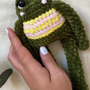 May include: A handmade green crochet frog plushie with large black eyes and a yellow and pink belly. The frog is made of a soft, textured yarn. A cute and cuddly stuffed animal.