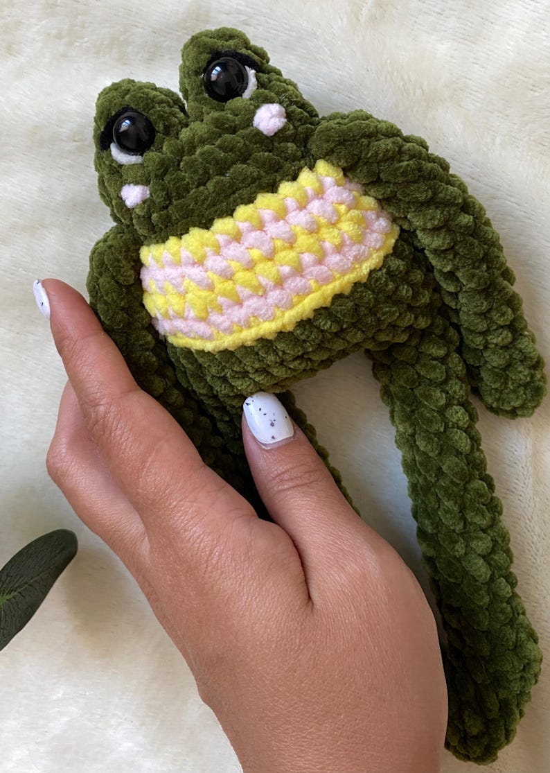 May include: A handmade green crochet frog plushie with large black eyes and a yellow and pink belly. The frog is made of a soft, textured yarn. A cute and cuddly stuffed animal.