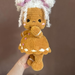 Puede incluir: Una mu&ntilde;eca de jengibre de crochet con pelo de lana blanca, lazos rosas y un vestido marr&oacute;n.