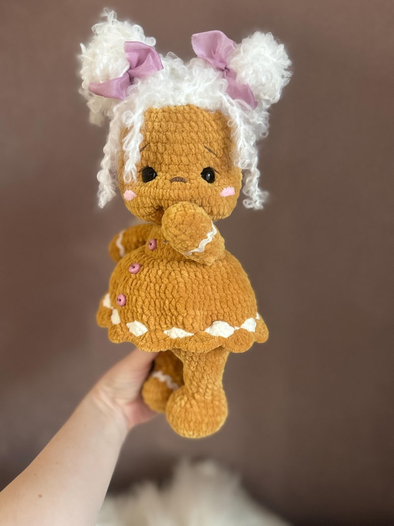 Puede incluir: Una mu&ntilde;eca de jengibre de crochet con pelo de lana blanca, lazos rosas y un vestido marr&oacute;n.