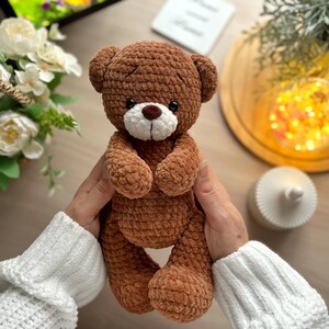 Puede incluir: Un oso de peluche marr&oacute;n hecho a mano con hocico blanco y nariz marr&oacute;n. El oso est&aacute; tejido con un hilo texturizado y tiene peque&ntilde;os ojos negros. El fondo incluye un arreglo floral y luces decorativas.