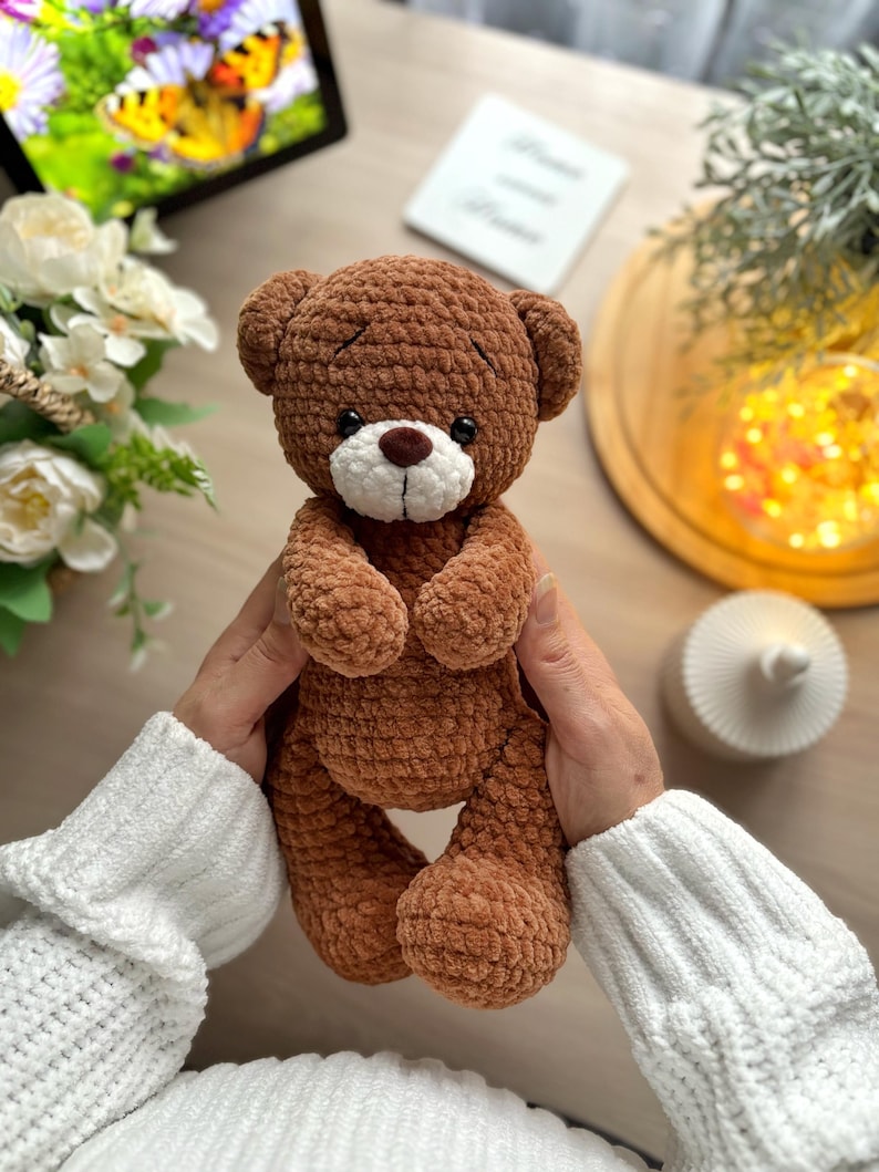 Puede incluir: Un oso de peluche marr&oacute;n hecho a mano con hocico blanco y nariz marr&oacute;n. El oso est&aacute; tejido con un hilo texturizado y tiene peque&ntilde;os ojos negros. El fondo incluye un arreglo floral y luces decorativas.