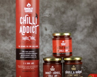 Op de afbeelding: Een set van drie rode glazen potten met Smokey Carter chili sauzen. De potten zijn gelabeld met "Scotch Bonnet Chili Jam", "Smokey Chipotle Chili Jam" en "Chipotle & Bourbon BBQ Sauce". De potten zijn in een piramidevorm gerangschikt op een grijze achtergrond. Een rode chilipeper bevindt zich op de voorgrond.