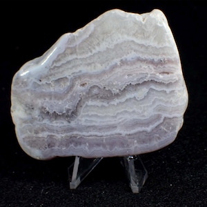 May include: A polished agate stone with layers of white, grey, and lavender colours. The natural stone is displayed on a clear acrylic stand, highlighting its unique patterns and textures. The background is black.