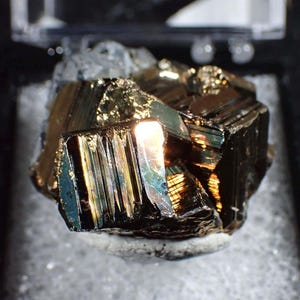 May include: A close-up of a pyrite mineral specimen. The metallic, golden-brown crystal has a reflective surface with iridescent blue and green hues. The mineral sits on a white, textured base, with a dark background.