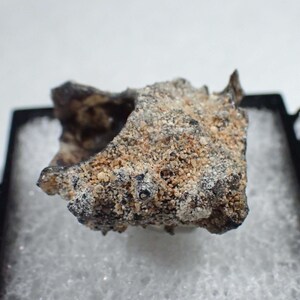 May include: A rough, irregular meteorite fragment featuring a textured surface with a mix of dark charcoal-gray and light tan, sandy-colored mineral deposits. The specimen is displayed resting on a white foam insert inside a black plastic case.