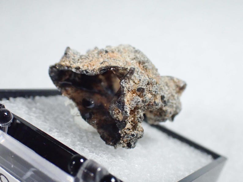 May include: A raw, irregular meteorite fragment with a dark, fused fusion crust and a porous, textured surface. The specimen displays a mix of charcoal black and sandy beige tones, showcasing a jagged, cratered exterior typical of space rock.