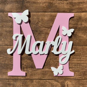 May include: Pink wooden letter M with the name "Marly" in white letters and white butterfly decorations.