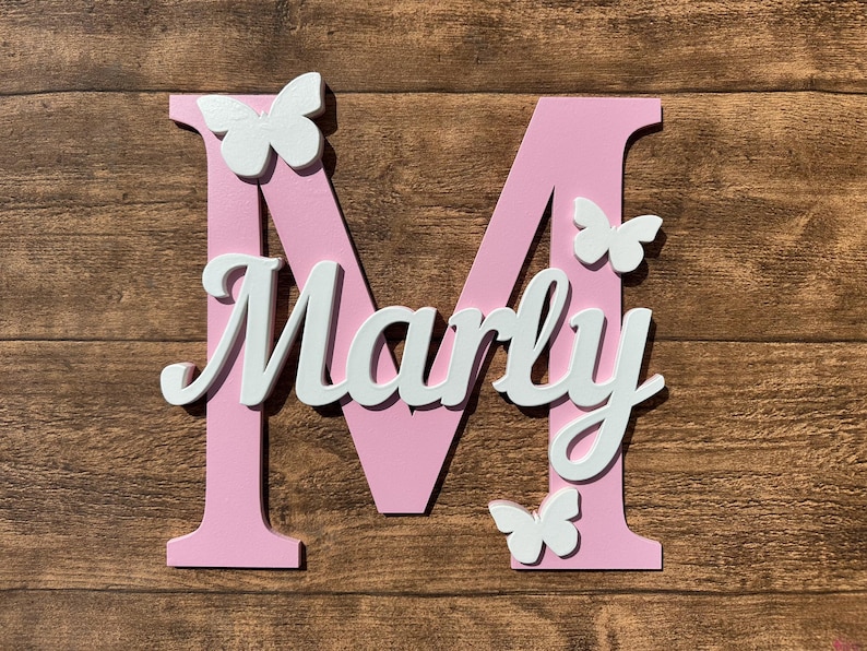 May include: Pink wooden letter M with the name "Marly" in white letters and white butterfly decorations.