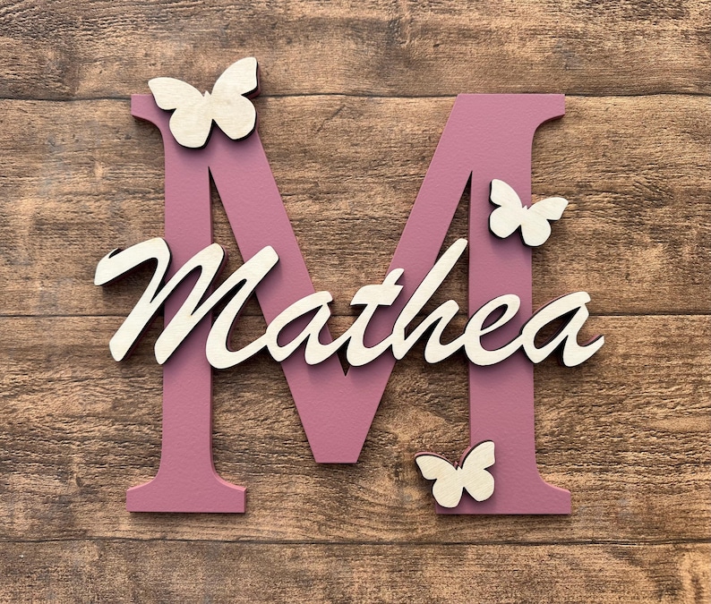May include: A pink wooden letter M with the name "Mathea" written in white on it. There are four white wooden butterflies attached to the letter and name.