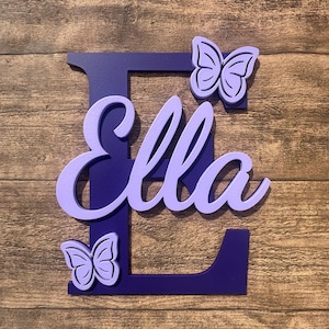 May include: Purple wooden letter E with the name "Ella" cut out in a lighter shade of purple. Two purple butterfly cutouts are on either side of the letter.