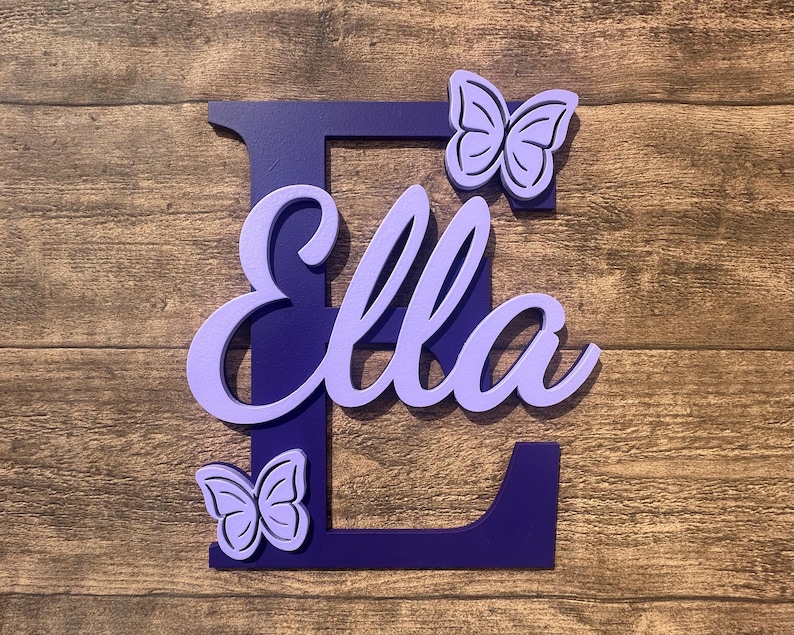 May include: Purple wooden letter E with the name "Ella" cut out in a lighter shade of purple. Two purple butterfly cutouts are on either side of the letter.