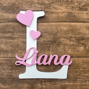 May include: White wooden letter L with the name Liana written in pink cursive letters. Three pink hearts are attached to the letter.