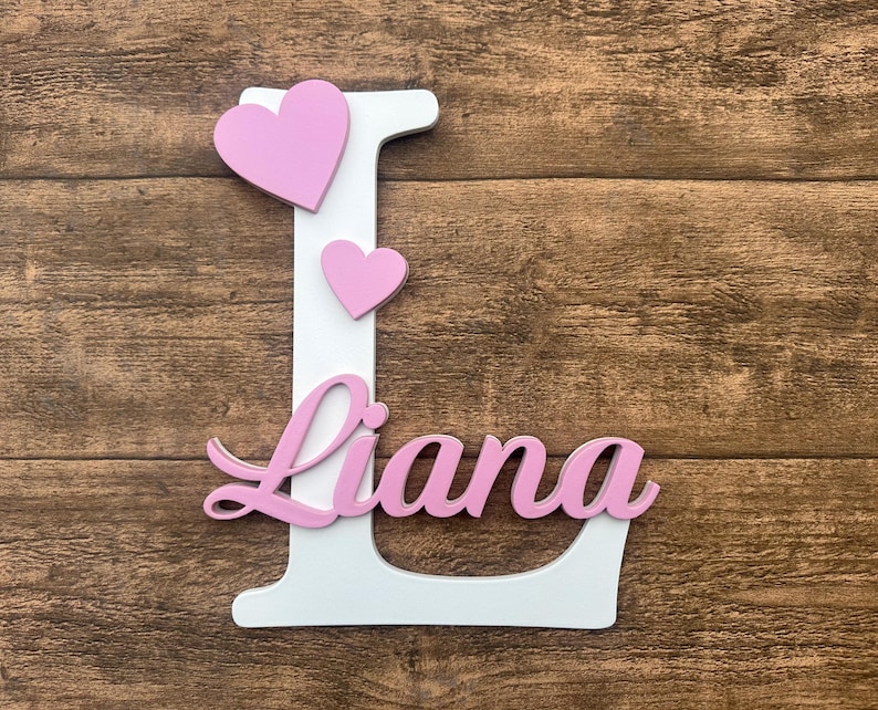 May include: White wooden letter L with the name Liana written in pink cursive letters. Three pink hearts are attached to the letter.