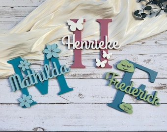 Op de afbeelding: Houten naamuitsparingen in teal, roze en groen. De namen "Mathilda", "Henrike" en "Frederick" worden weergegeven met decoratieve elementen zoals vlinders en bloemen. De letters zijn ongeveer 15 cm hoog.