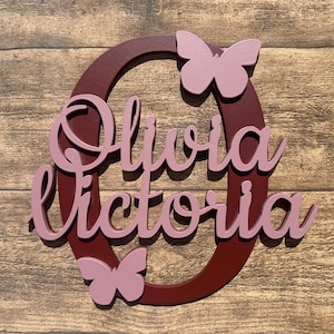 May include: A burgundy wooden circle with the names Olivia Victoria written in pink cursive letters. Two pink butterflies are attached to the circle.