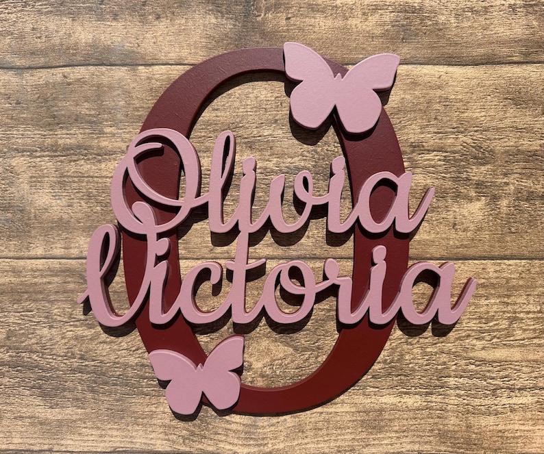 May include: A burgundy wooden circle with the names Olivia Victoria written in pink cursive letters. Two pink butterflies are attached to the circle.