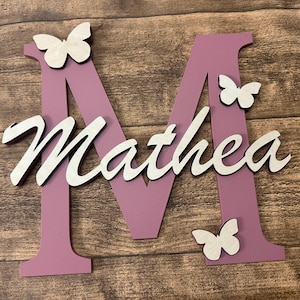 May include: A pink wooden letter M with the name Mathea written in white on it. There are three white butterflies on the letter and the background is a wooden surface.