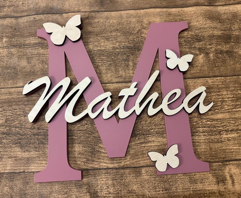 May include: A pink wooden letter M with the name Mathea written in white on it. There are three white butterflies on the letter and the background is a wooden surface.